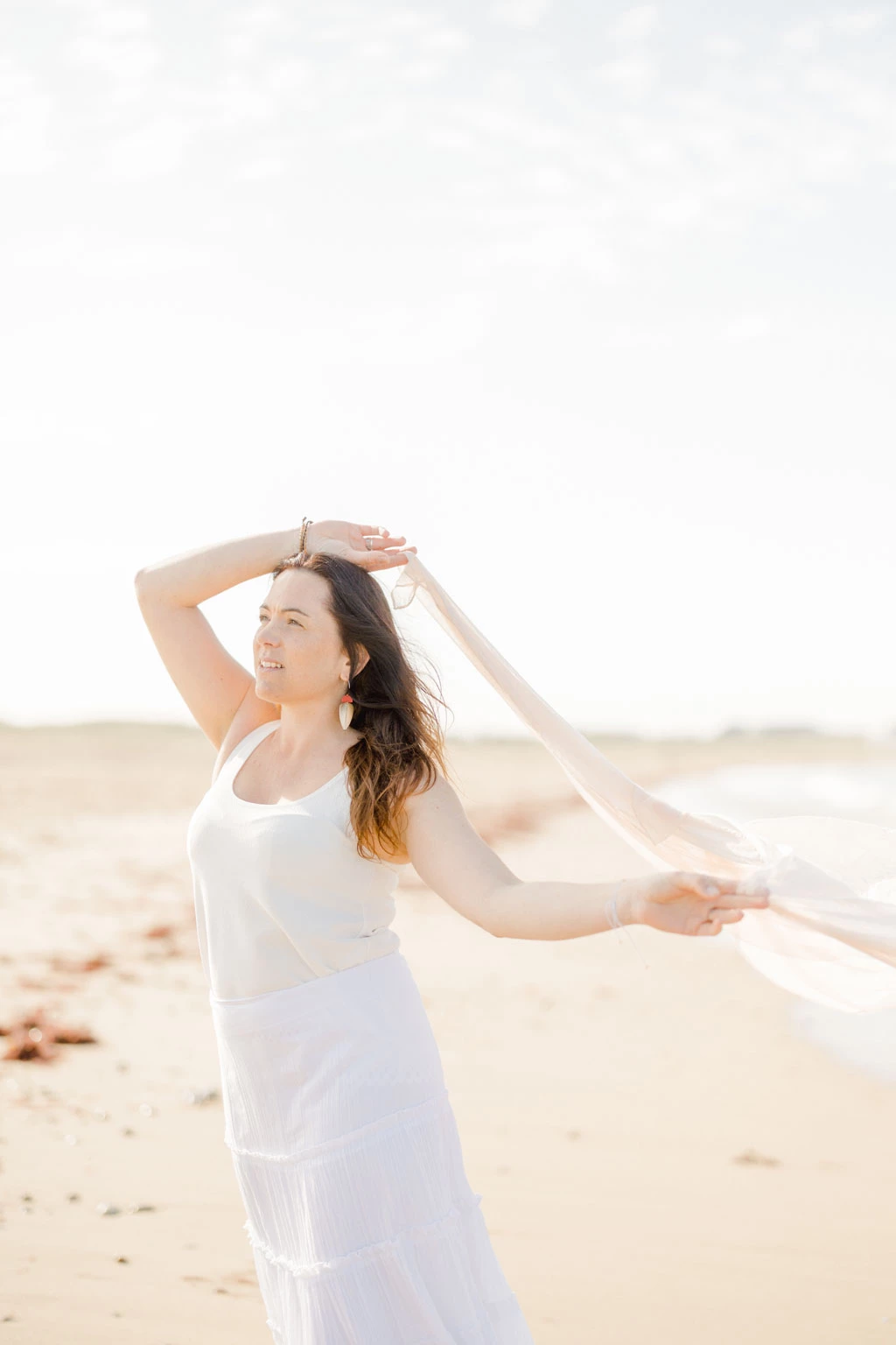 Femme en robe blanche sur une plage ensoleillée, levant les bras dans un geste de liberté et de joie
