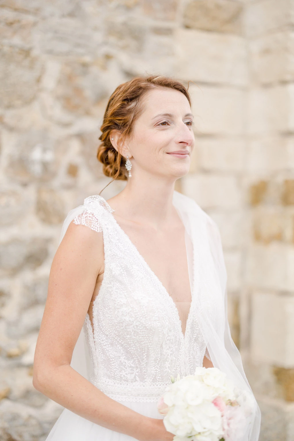 Mariée souriante en robe blanche avec bouquet, regard levé vers le ciel dans un décor de pierre