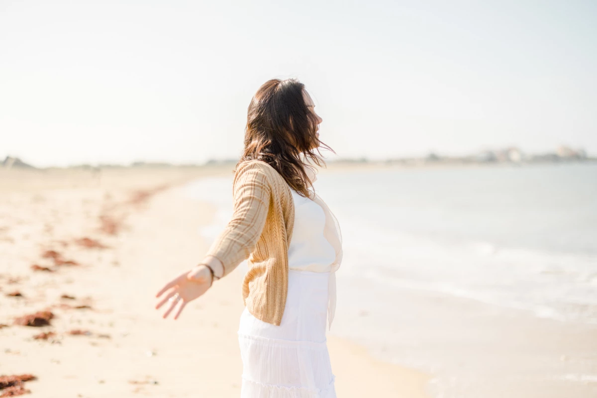 Femme enceinte en robe blanche les bras ouverts sur une plage de sable au bord de l'eau