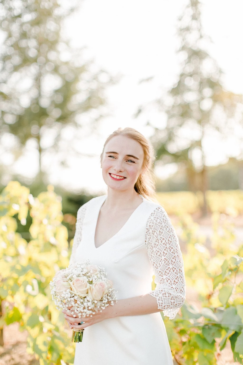 Mariée souriante en robe blanche tenant un bouquet de gypsophile dans un vignoble au coucher du soleil