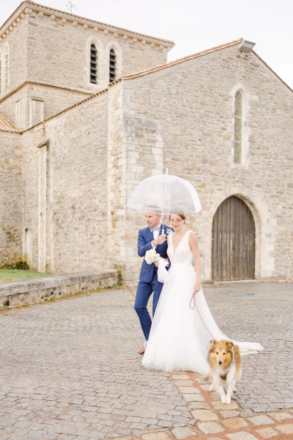 Couple de mariés avec leur chien devant une église en pierre sous un parapluie blanc