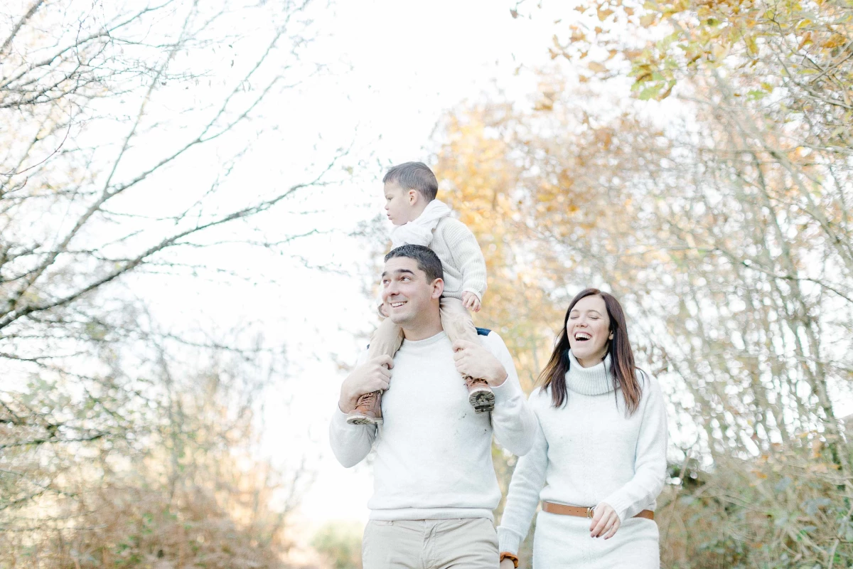 Famille complice en tenue blanche se promenant dans la nature automnale avec leur enfant sur les épaules