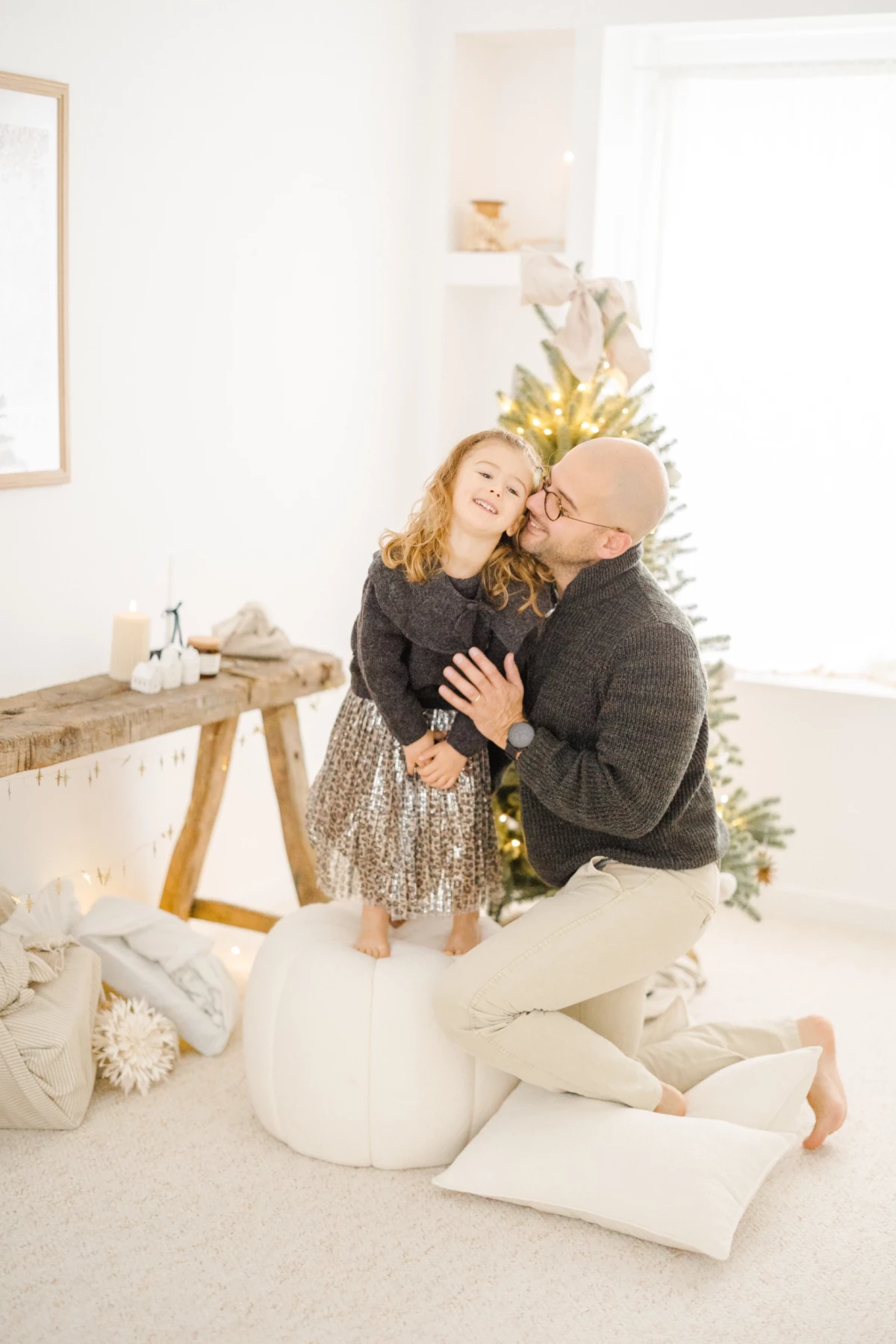 Père et fille enlacés devant un sapin de Noël dans un studio lumineux aux tons blanc et beige