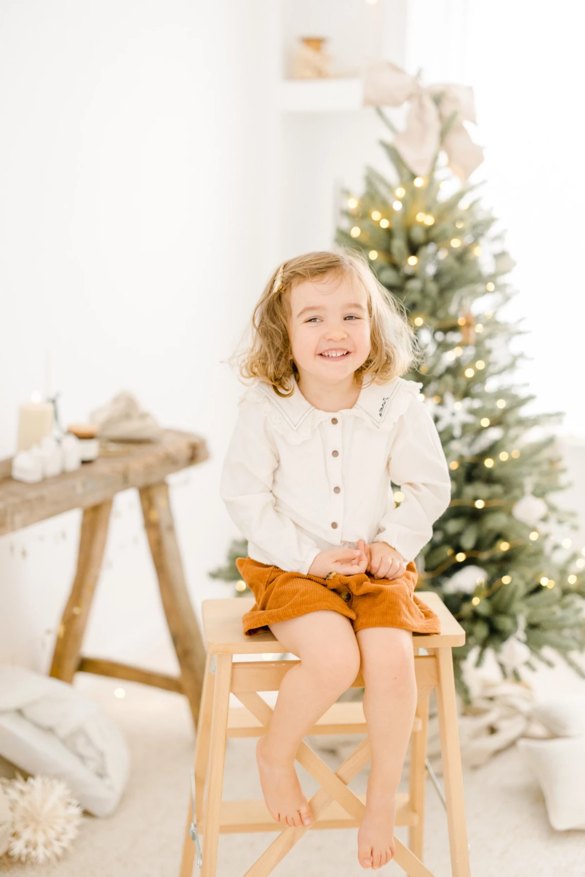 Petite fille souriante assise sur un tabouret en bois devant un sapin de Noël illuminé