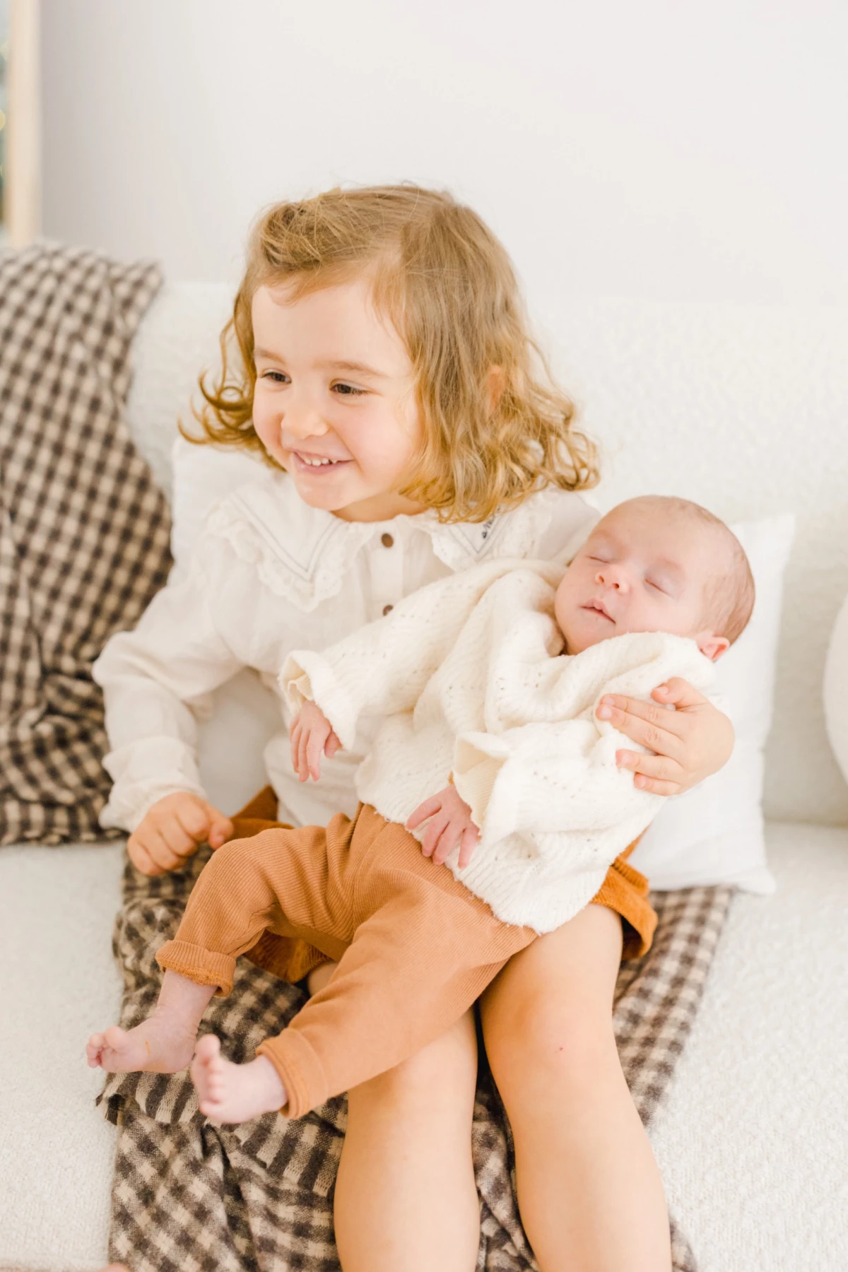 Grande sœur souriante tenant tendrement son nouveau-né dans les bras sur un canapé à carreaux