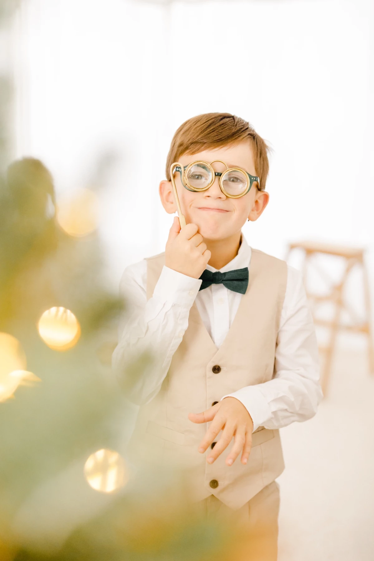 Jeune garçon en costume beige et nœud papillon, lunettes rondes, geste pensif lors de préparatifs de mariage