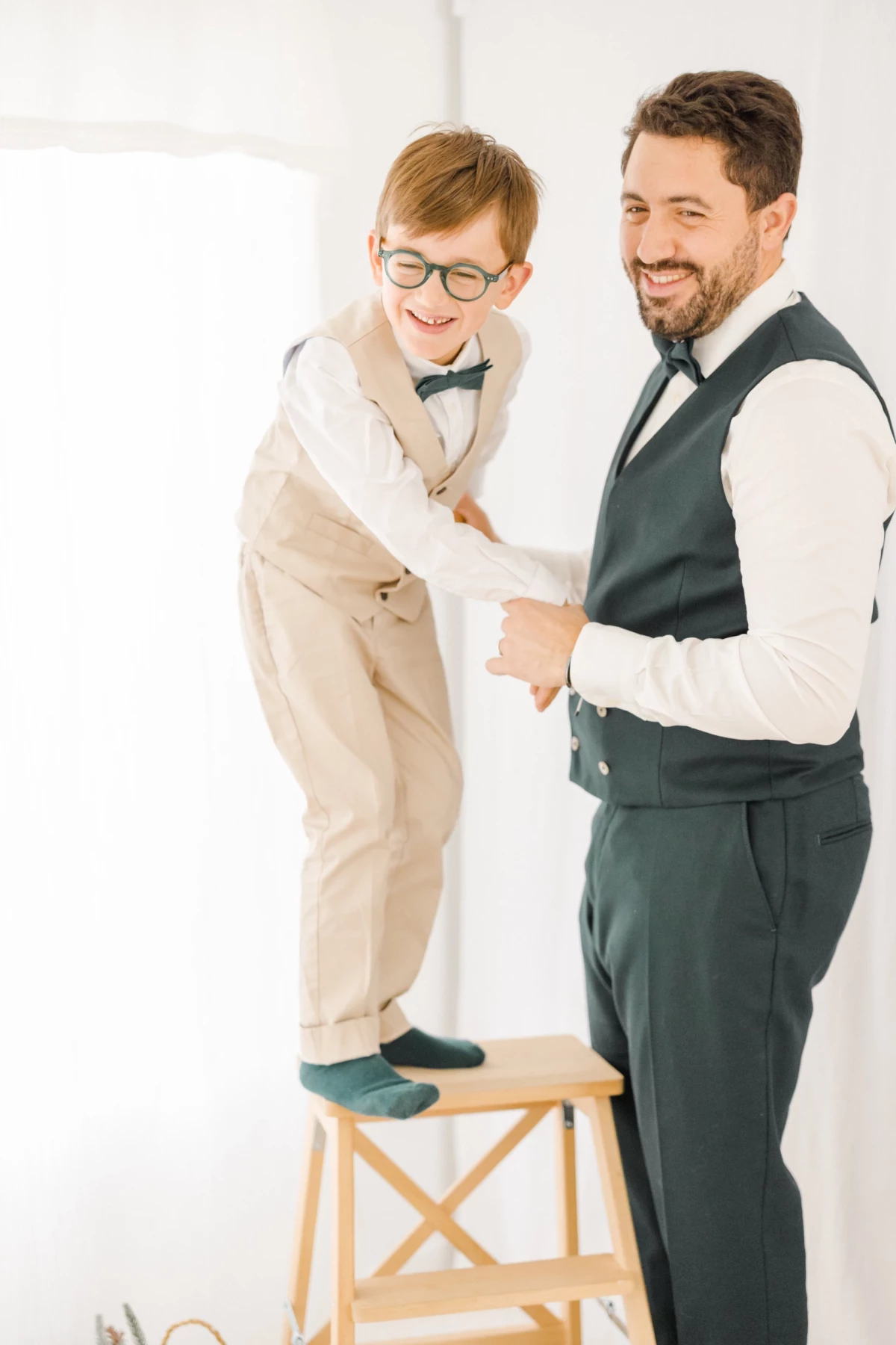 Père et fils complices en costume lors des préparatifs de mariage dans un studio lumineux