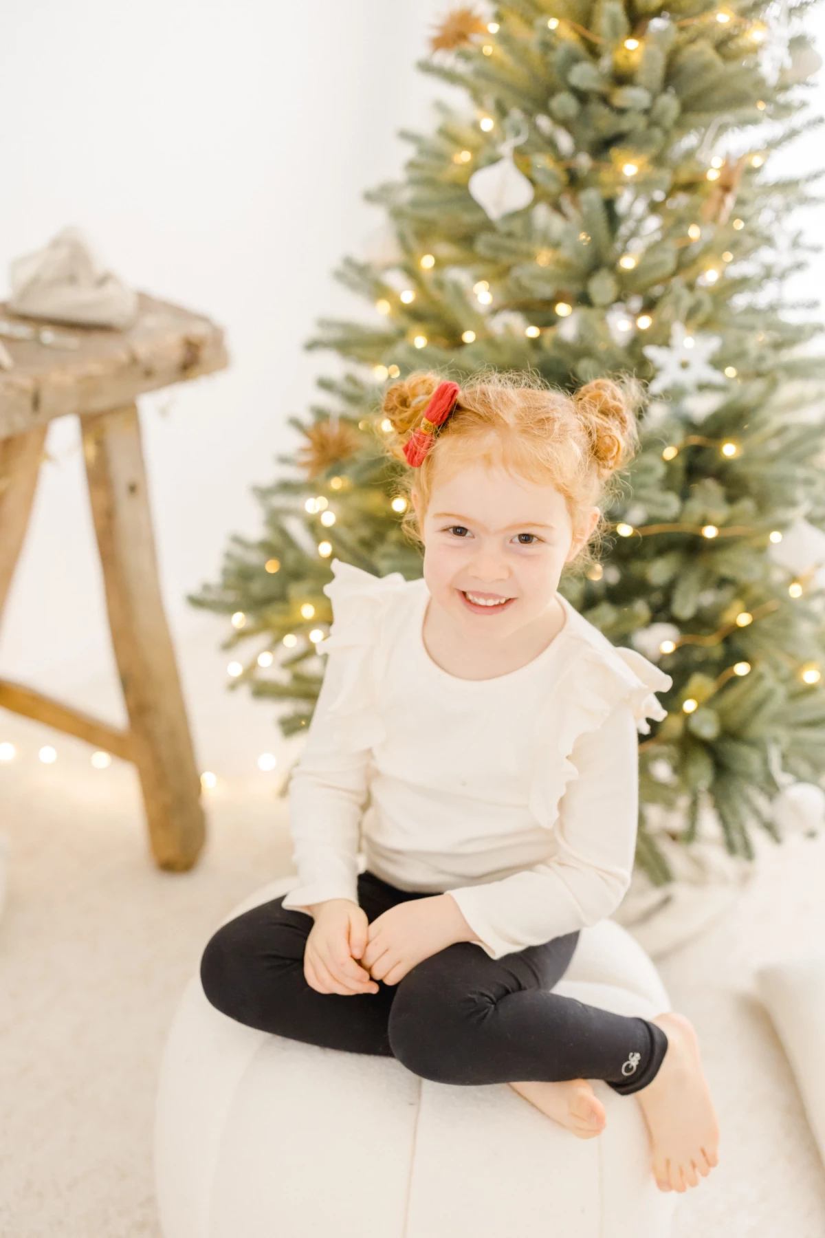 Petite fille souriante assise devant un sapin de Noël illuminé, vêtue d'un pull blanc
