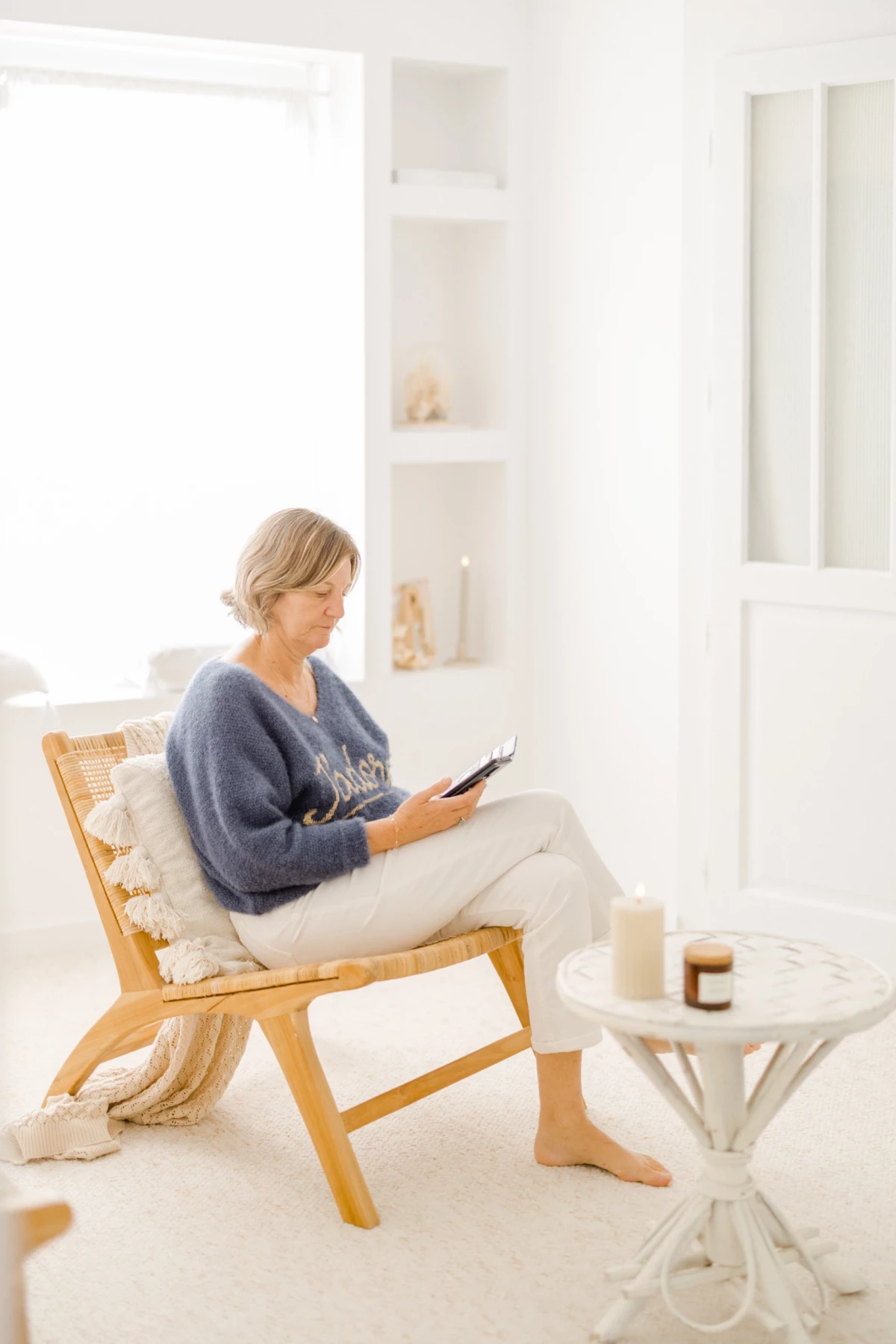 Femme en pull confortable lisant paisiblement dans un fauteuil en bois dans un intérieur blanc épuré