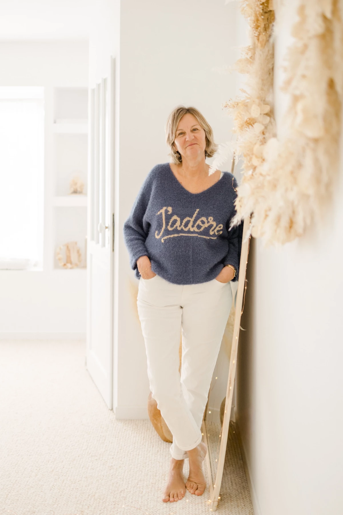 Portrait professionnel d'une femme souriante en pull bleu marine, adossée à un mur blanc décoré de pampas