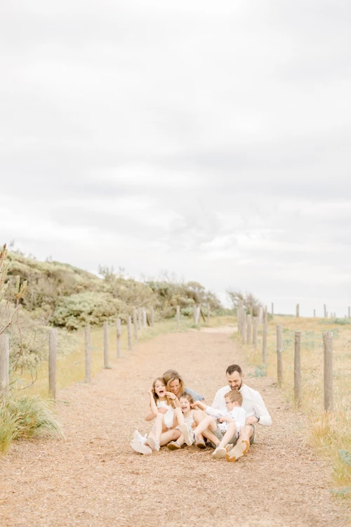 Famille heureuse assise sur un chemin de sable bordé de dunes, vêtue de blanc dans une ambiance douce