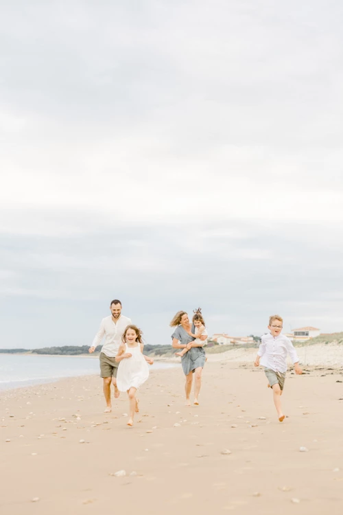Famille courant pieds nus sur une plage de sable fin sous un ciel voilé, parents et enfants main dans la main