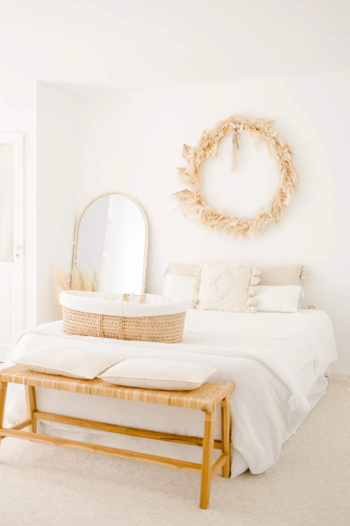 Chambre de bébé épurée avec berceau en osier, miroir ovale et couronne de fleurs séchées sur mur blanc