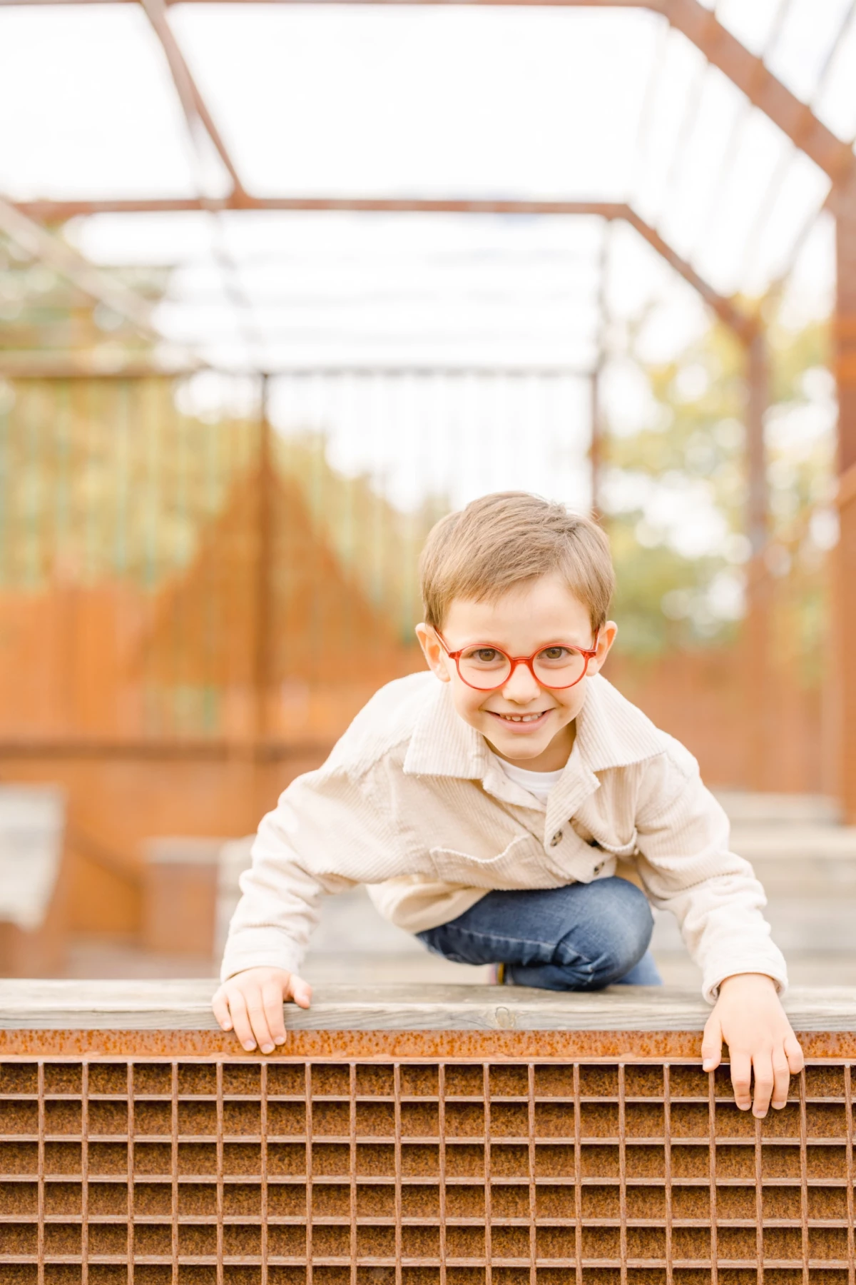 Jeune garçon souriant avec lunettes rouges, assis sur une structure métallique dorée en extérieur