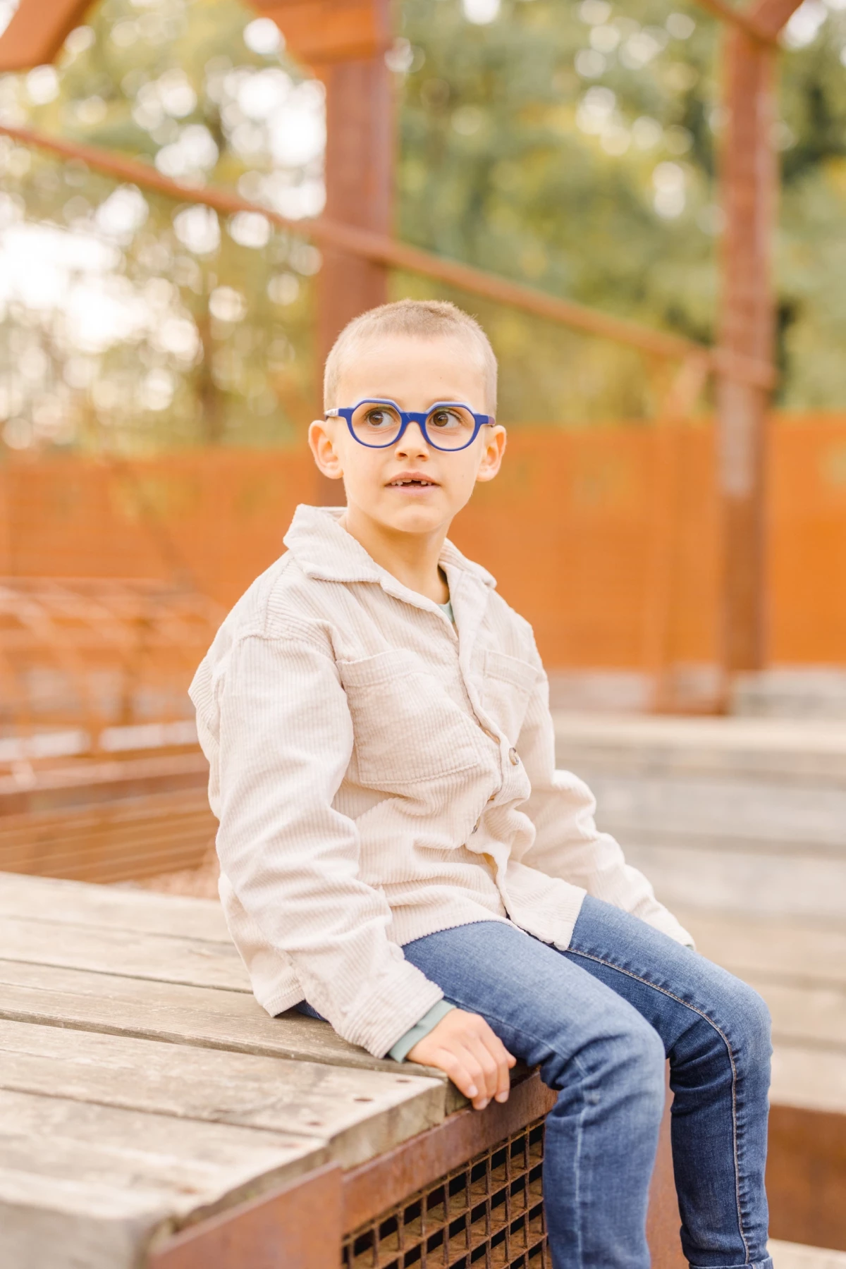 Portrait d'un jeune garçon blond à lunettes souriant, assis sur un banc en bois en extérieur