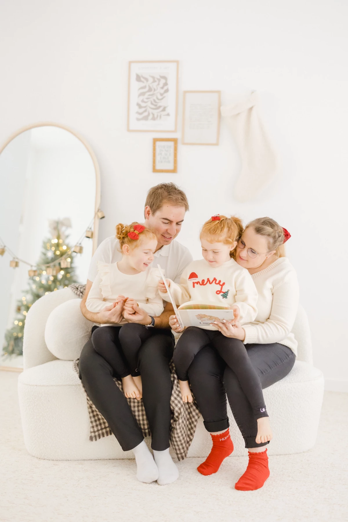 Mère et ses trois enfants lisant ensemble un livre sur un canapé blanc dans un décor de Noël lumineux