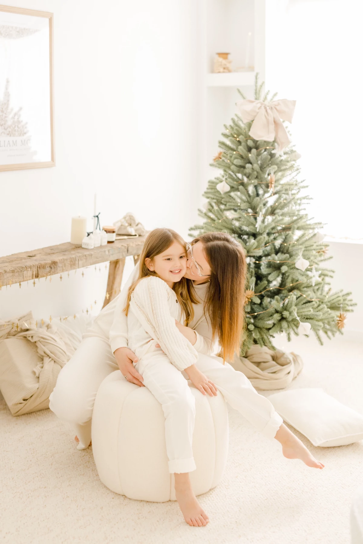 Mère et fille complices en tenue blanche devant un sapin de Noël enneigé dans un décor chaleureux