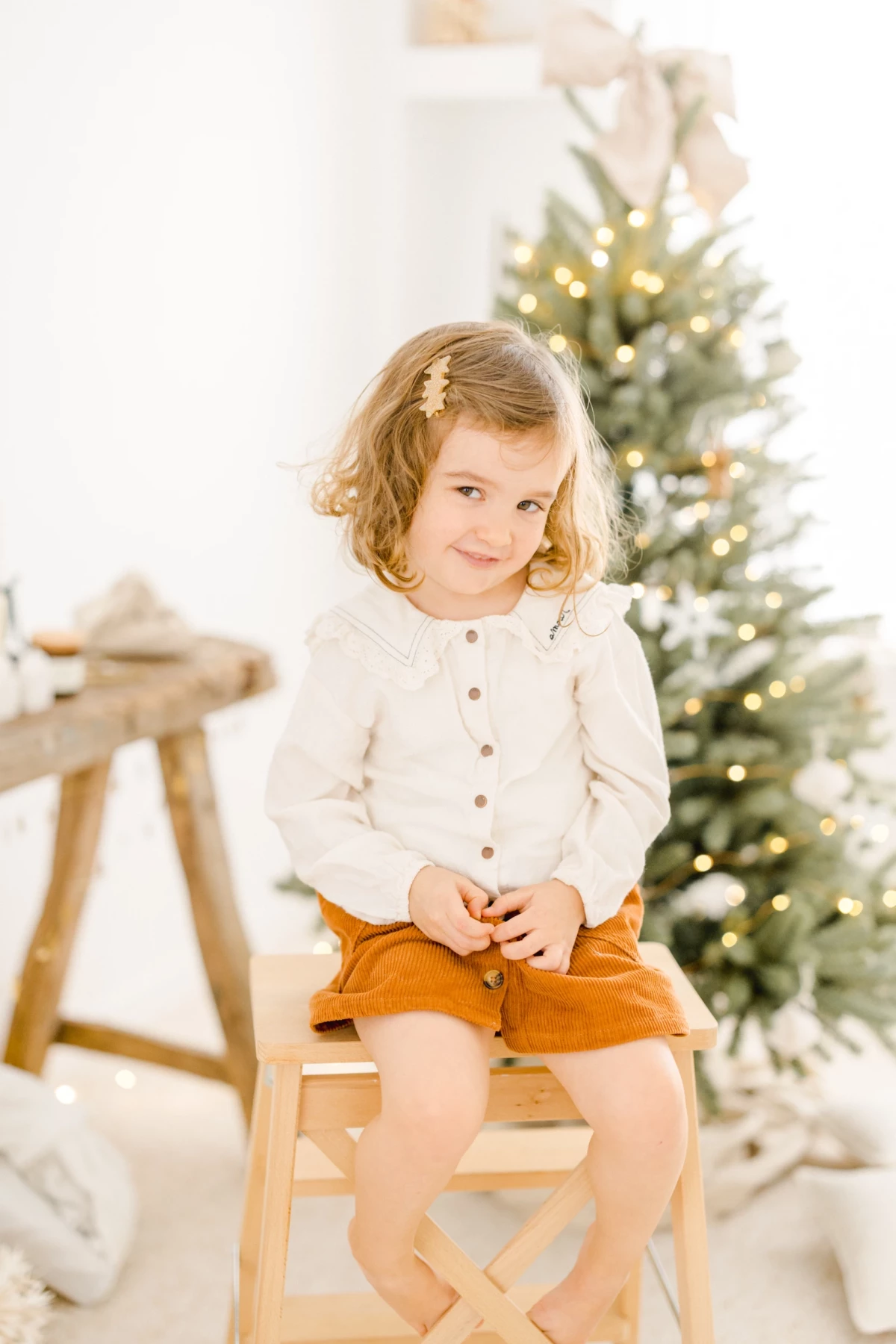 Petite fille souriante assise devant un sapin de Noël illuminé, portant une tenue blanche et un short camel
