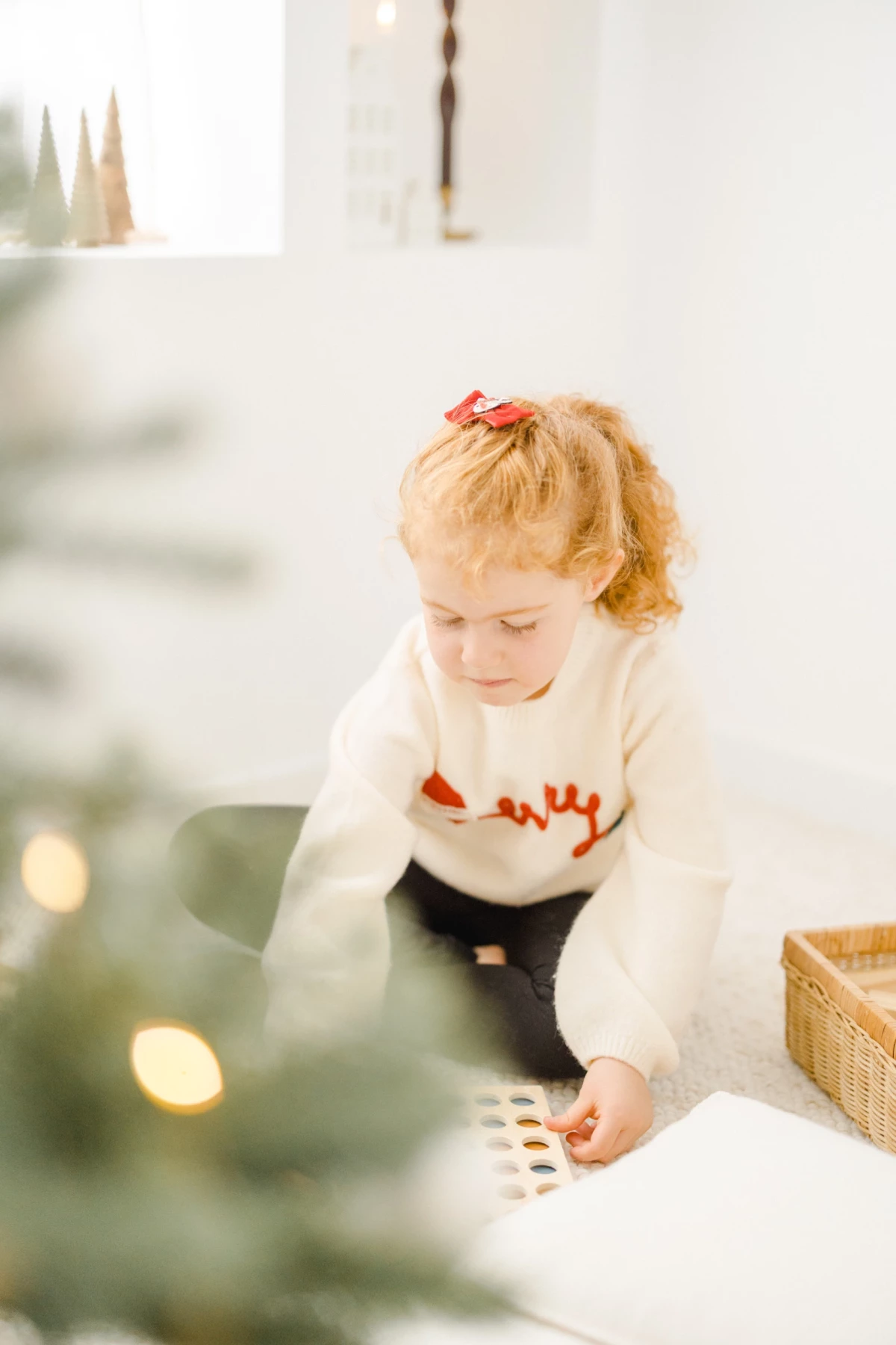 Petite fille blonde en pull blanc jouant près du sapin de Noël illuminé dans une ambiance chaleureuse