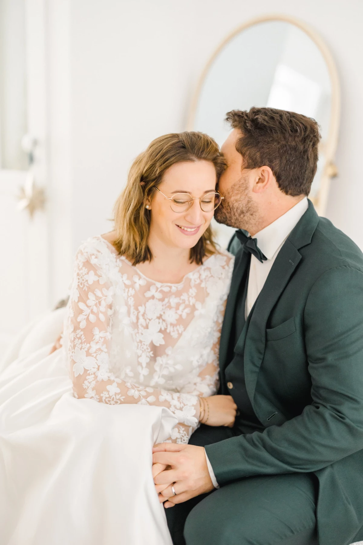 Couple de mariés assis tendrement, lui en costume sombre embrassant sa joue, elle en robe blanche fleurie