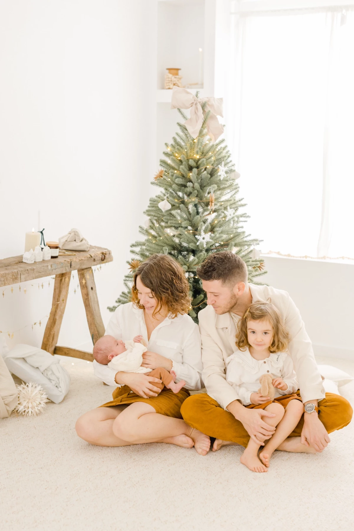 Famille réunie au sol devant un sapin de Noël, parents et deux enfants en tenues coordonnées blanches et ocre