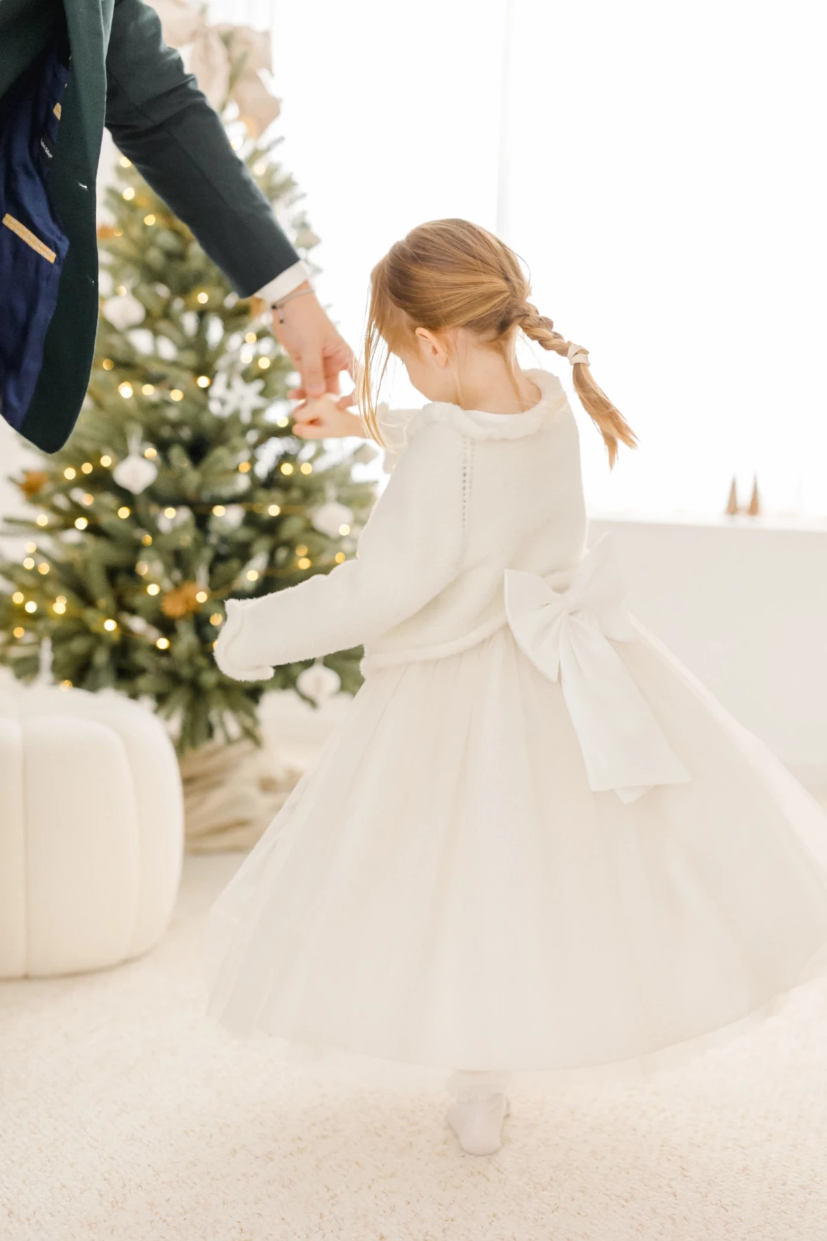 Petite fille en robe blanche tournoyant près d'un sapin de Noël illuminé en tendant la main vers un adulte