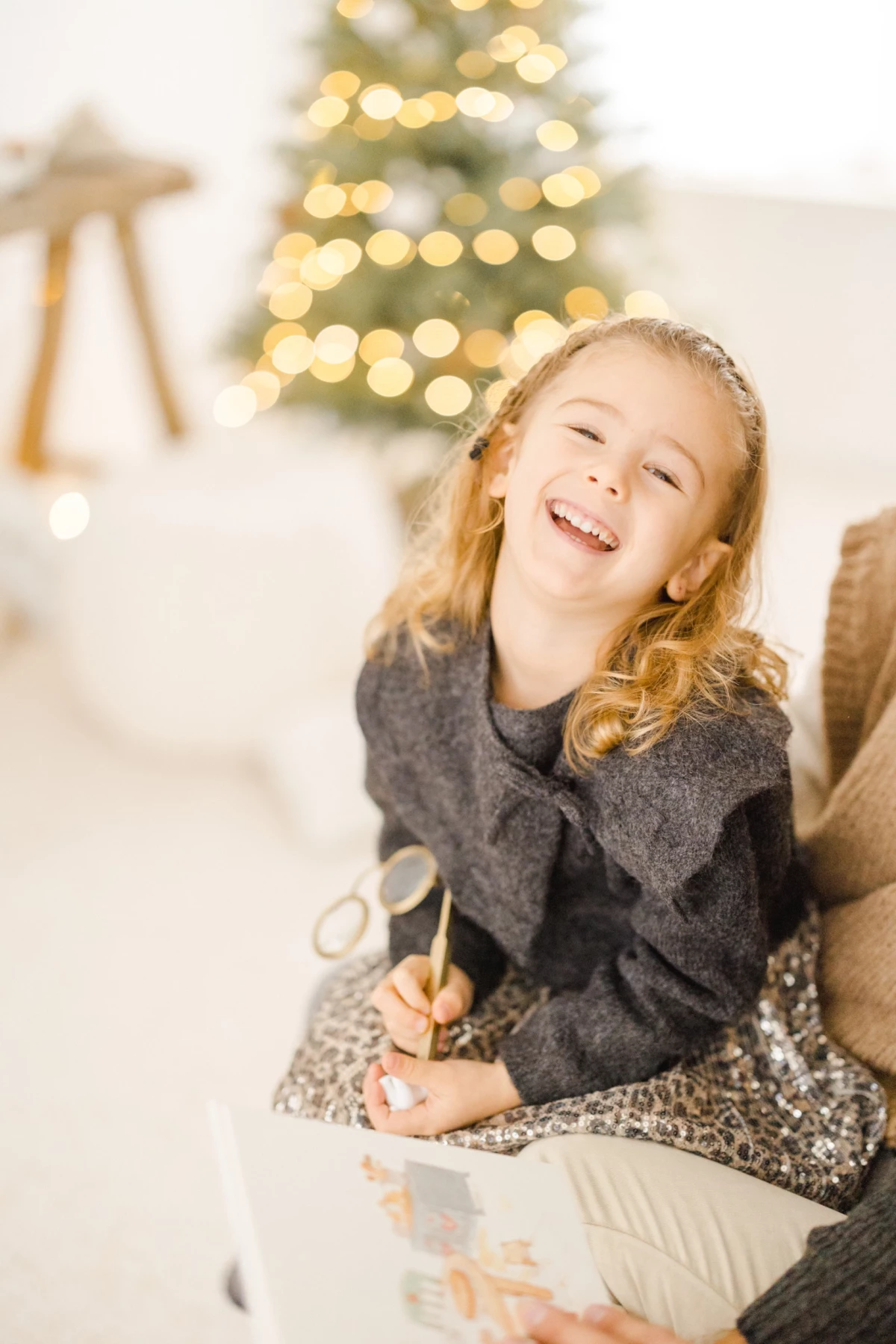 Petite fille souriante en pull gris tenant des ciseaux dorés devant un sapin de Noël illuminé