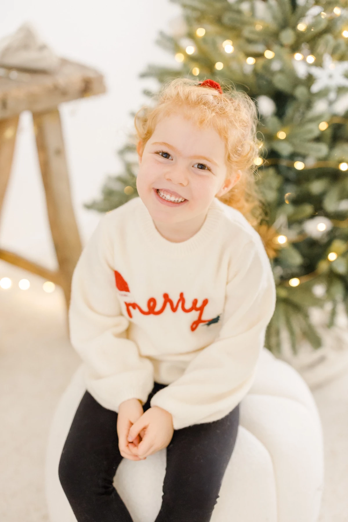 Petite fille souriante en pull blanc devant un sapin de Noël illuminé