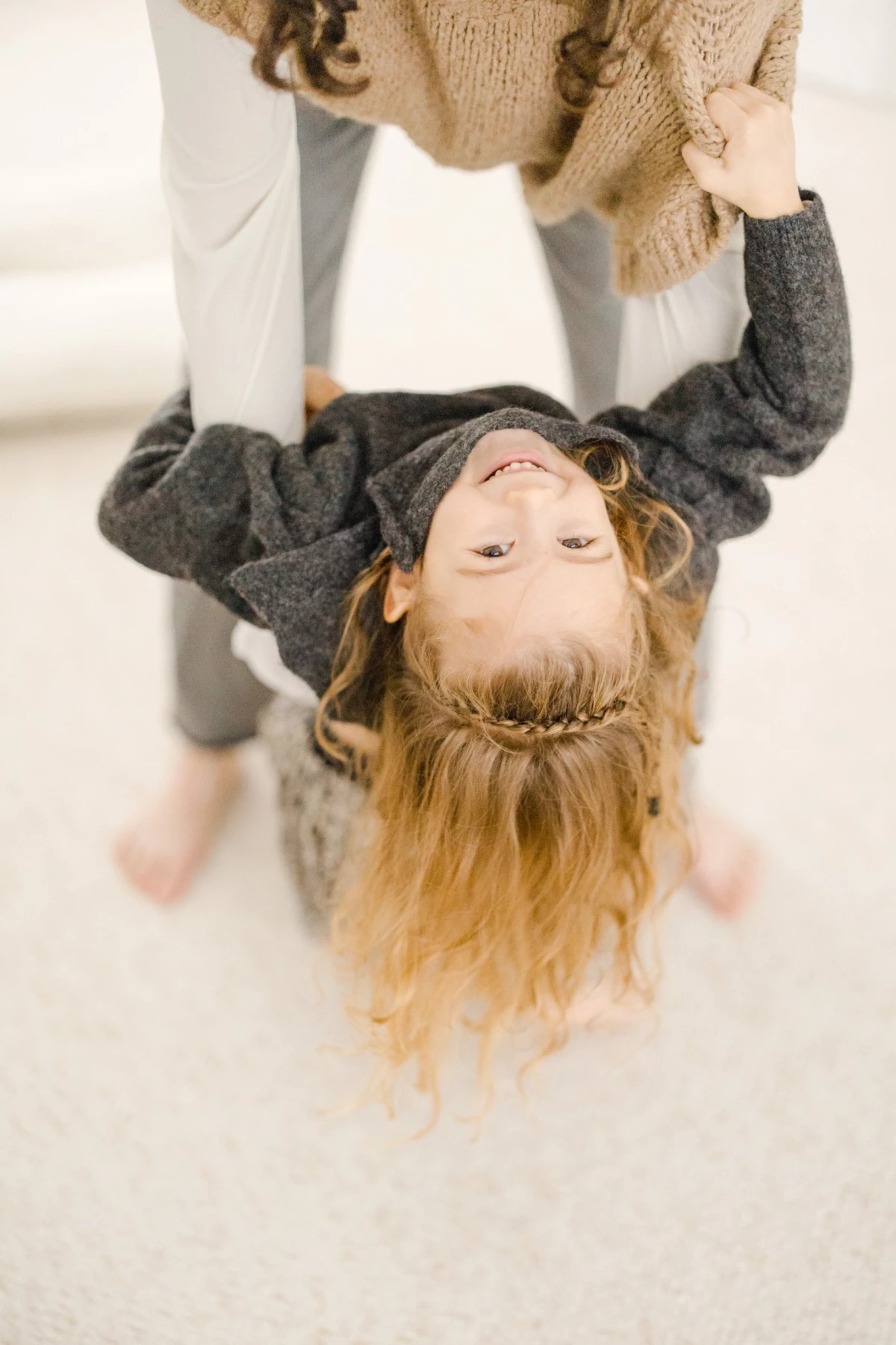 Petite fille aux cheveux bouclés suspendue à l'envers, sourire éclatant, tenue par les mains d'un adulte