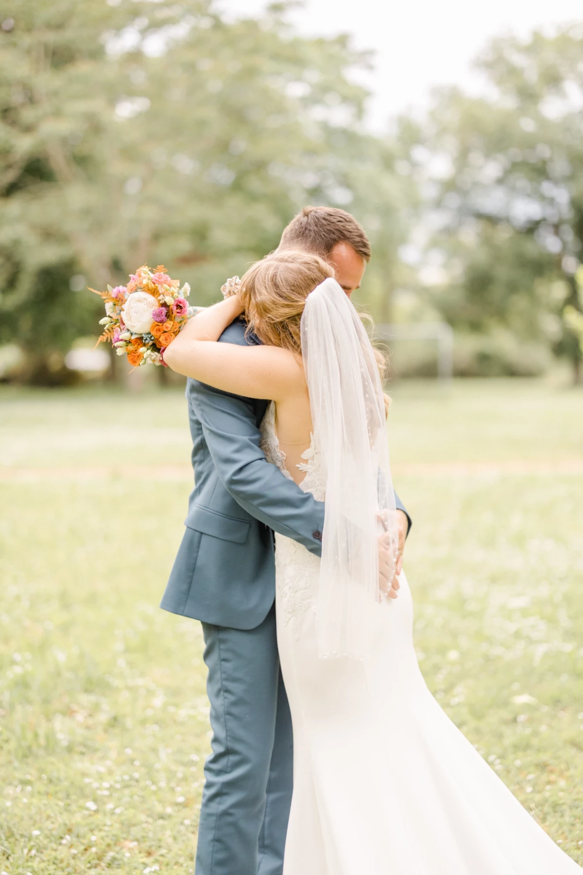 Couple de mariés s'enlaçant tendrement dans un parc verdoyant, la mariée tient un bouquet orange