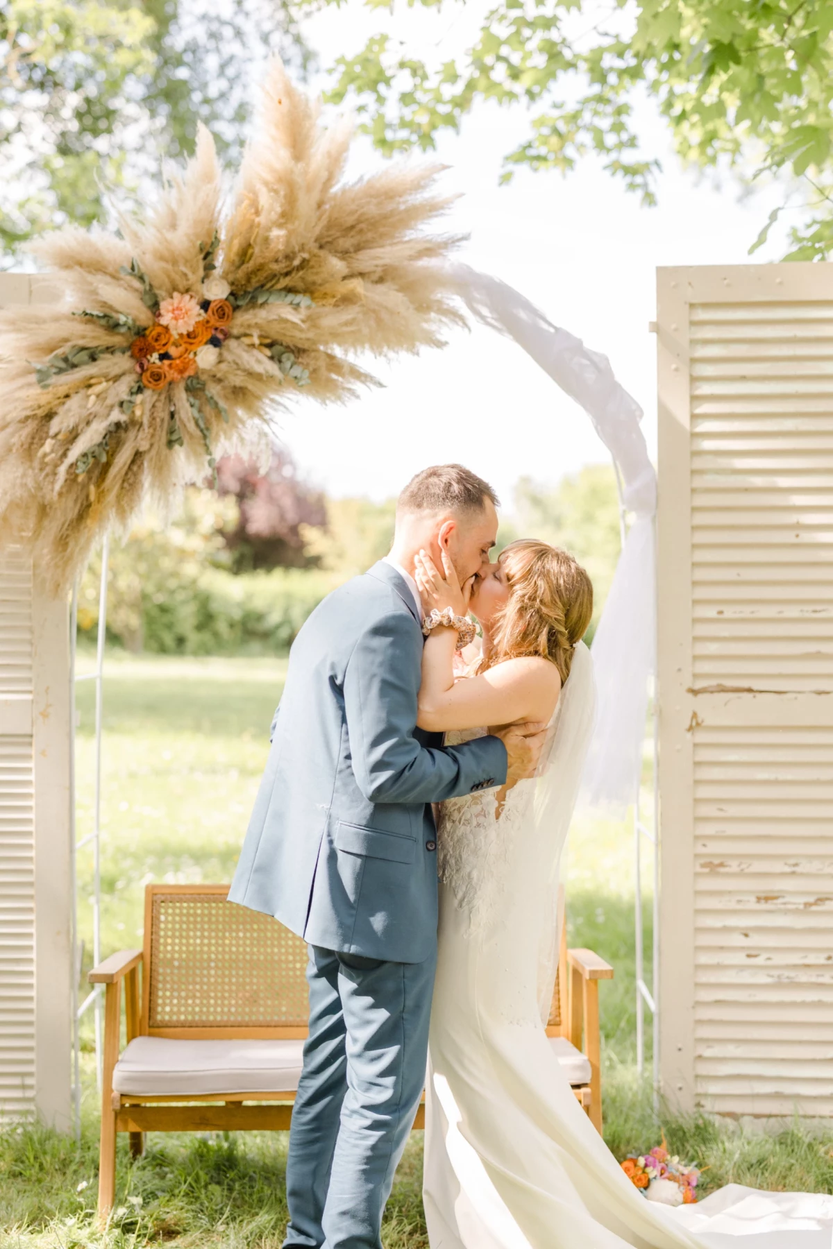 Couple de mariés s'embrassant sous un arc décoré de pampas beige et fleurs orangées en extérieur