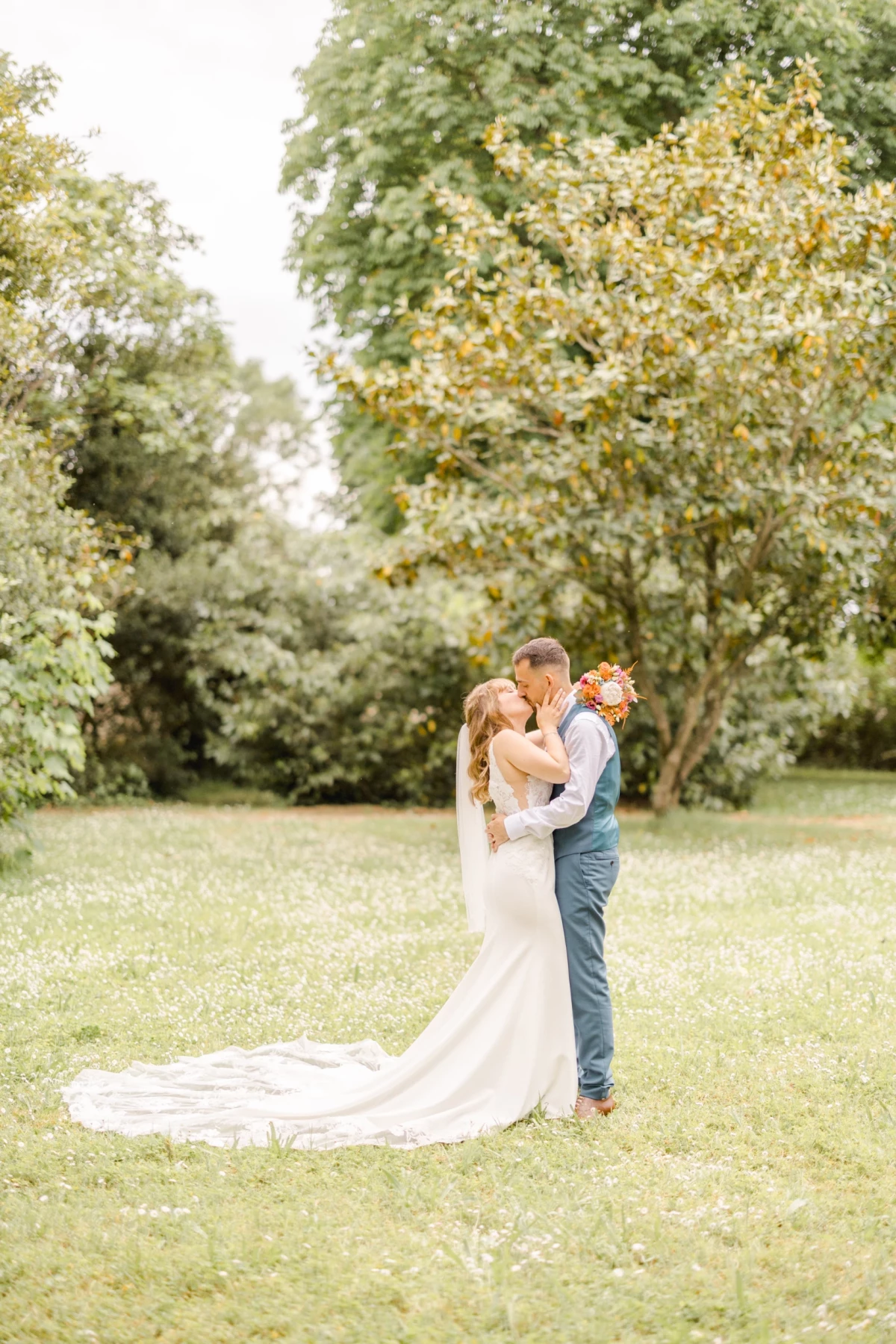 Couple de mariés s'embrassant dans un parc verdoyant, robe blanche à longue traîne