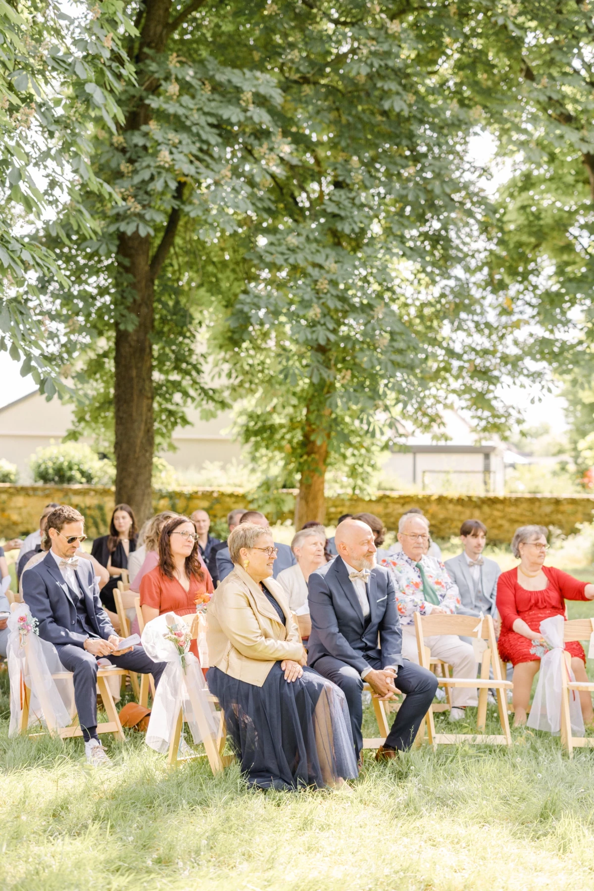 Invités de mariage assis sur des chaises blanches sous de grands arbres lors d'une cérémonie en extérieur