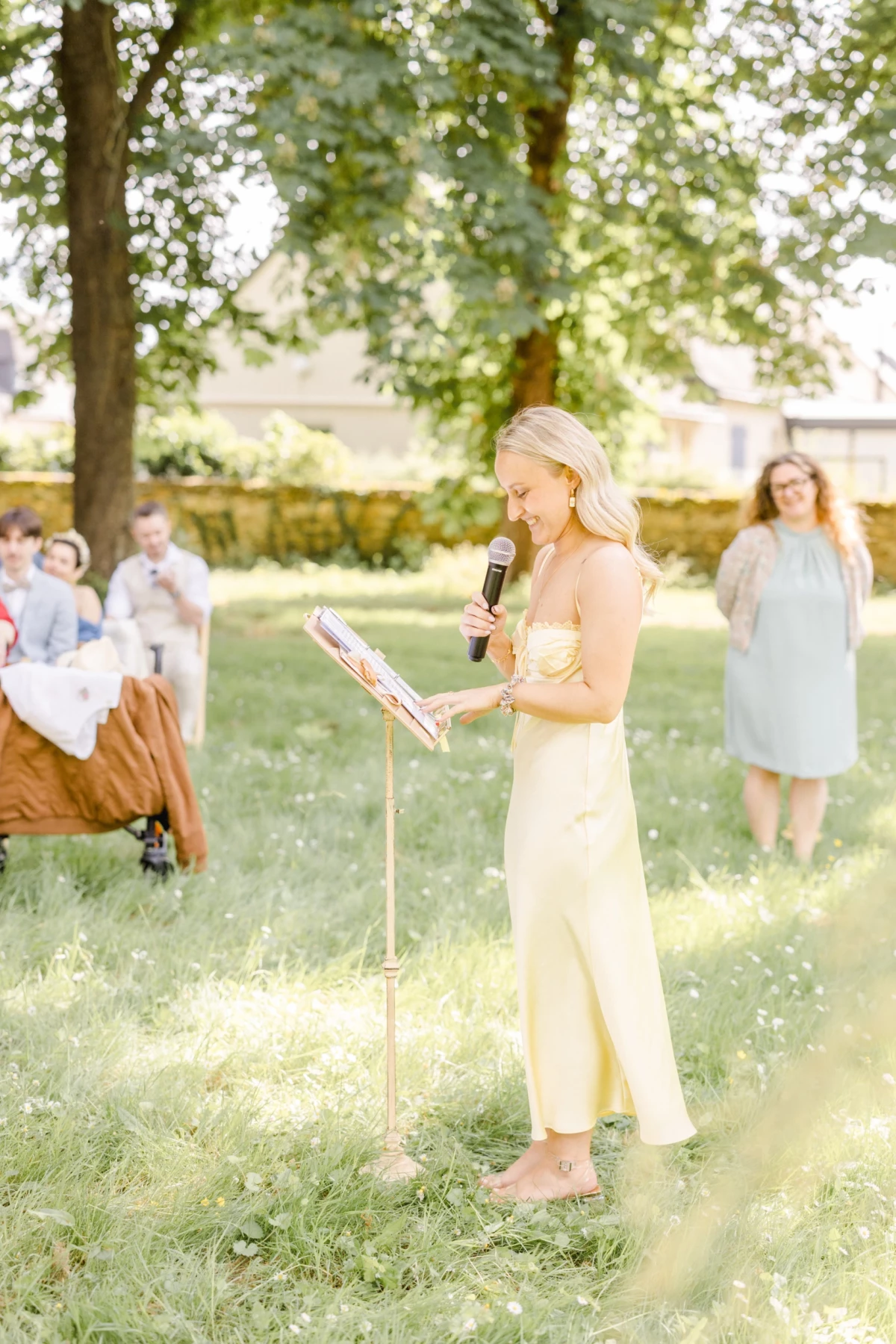 Officiant de cérémonie laïque en robe jaune lisant un texte face aux invités sous les arbres