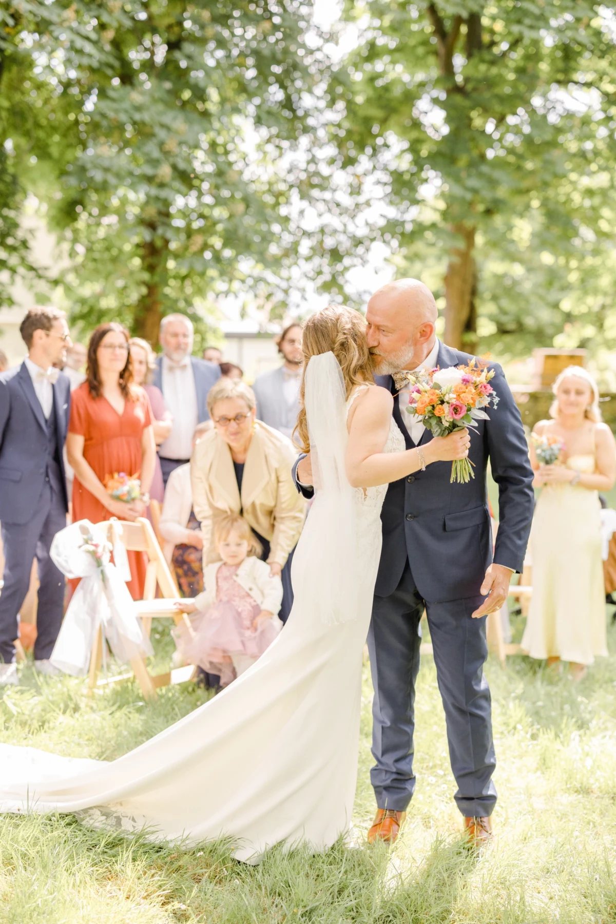 Couple de mariés s'embrassant lors d'une cérémonie en plein air sous les arbres avec leurs invités