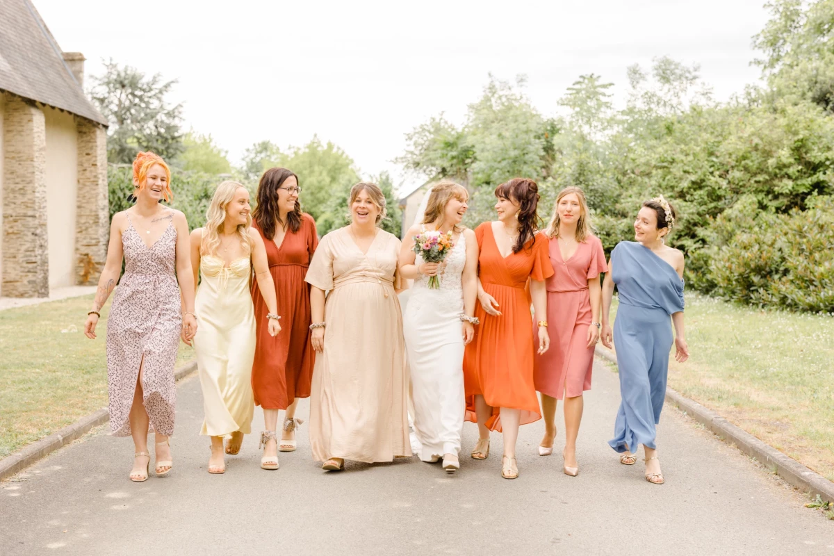Cortège de mariage en robes colorées marchant dans une allée ensoleillée, entourant la mariée en robe blanche