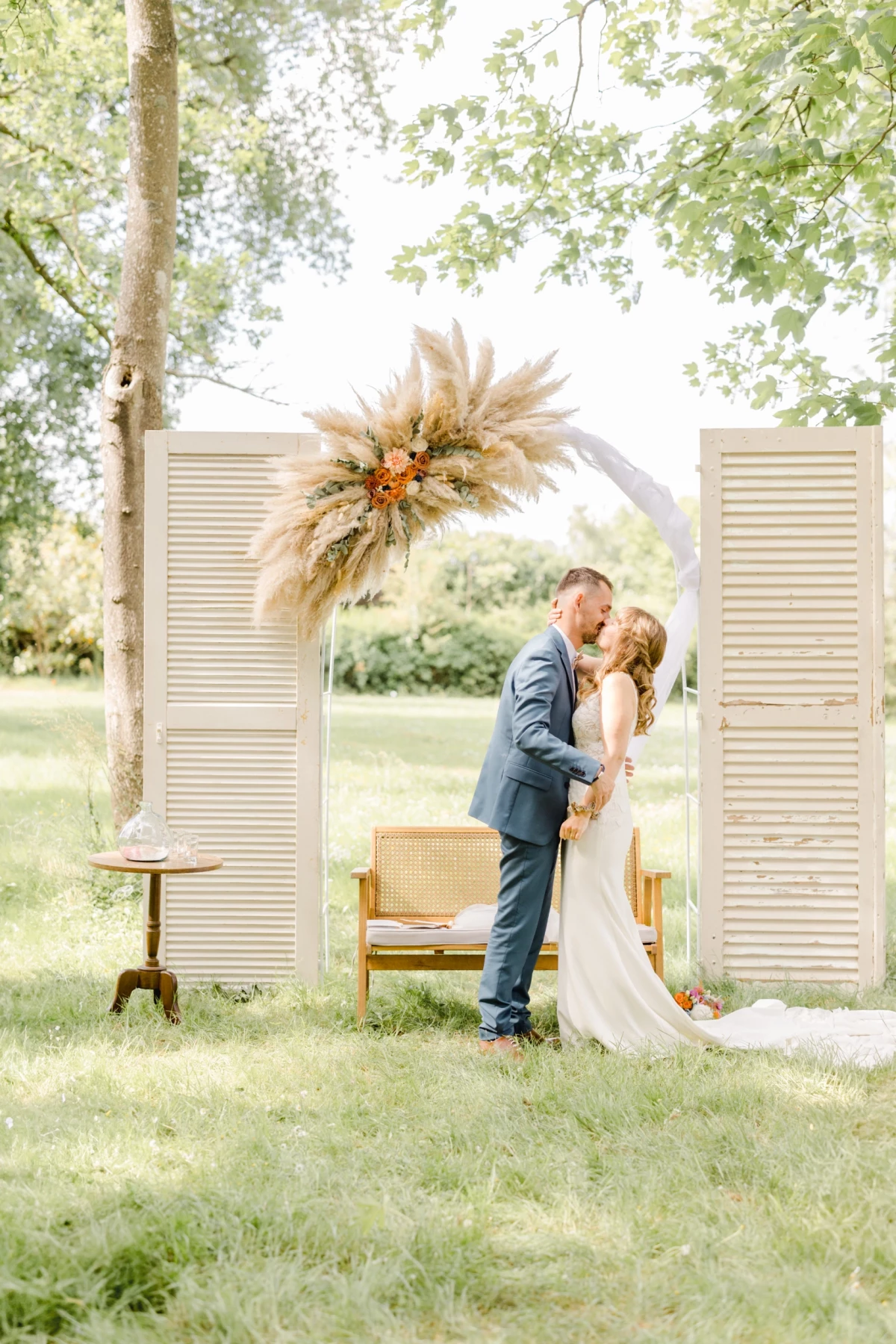Couple de mariés s'embrassant devant des volets blancs et une arche de fleurs séchées en extérieur
