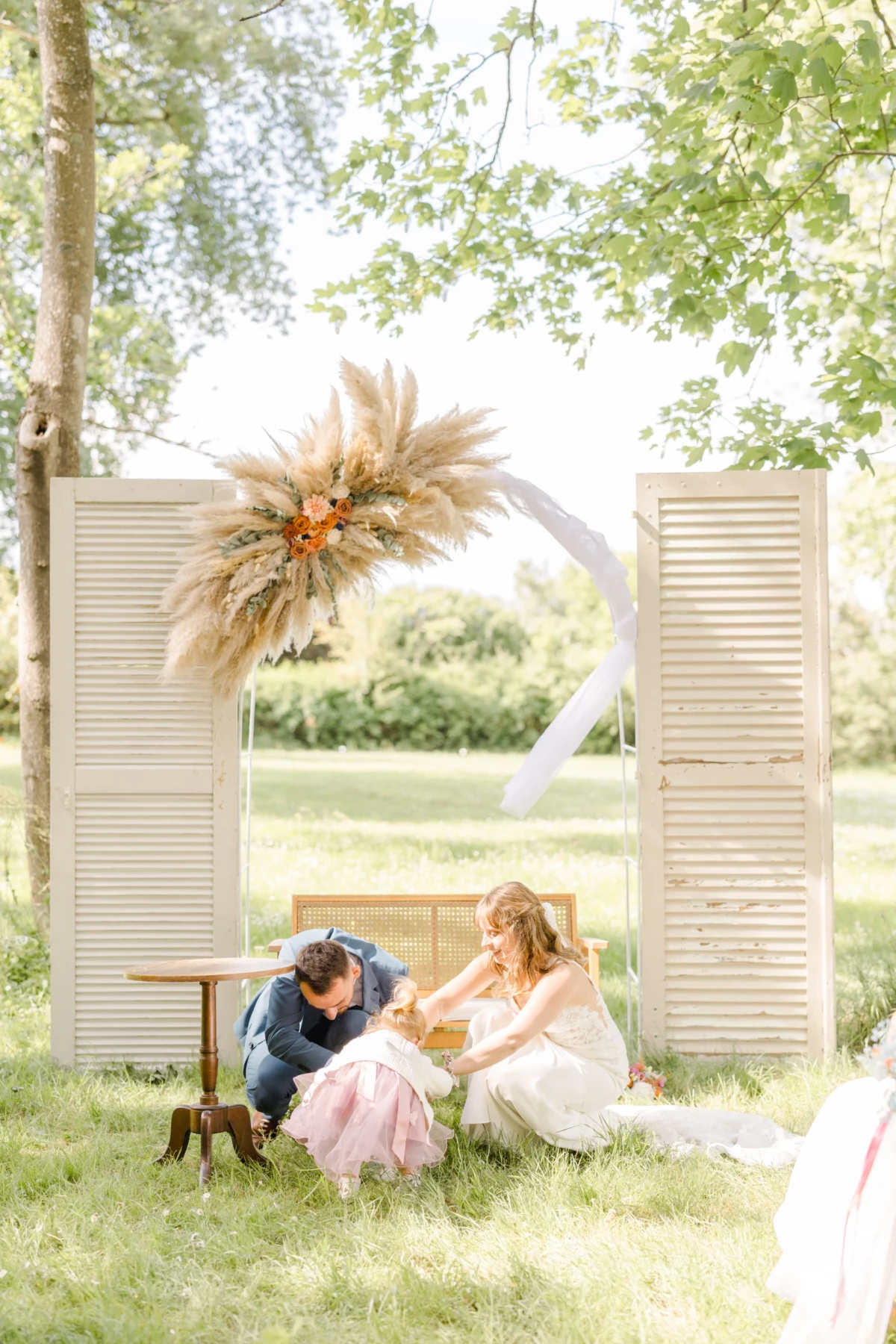 Couple de mariés assis sur un banc vintage entre portes blanches sous arche florale de pampa séchée