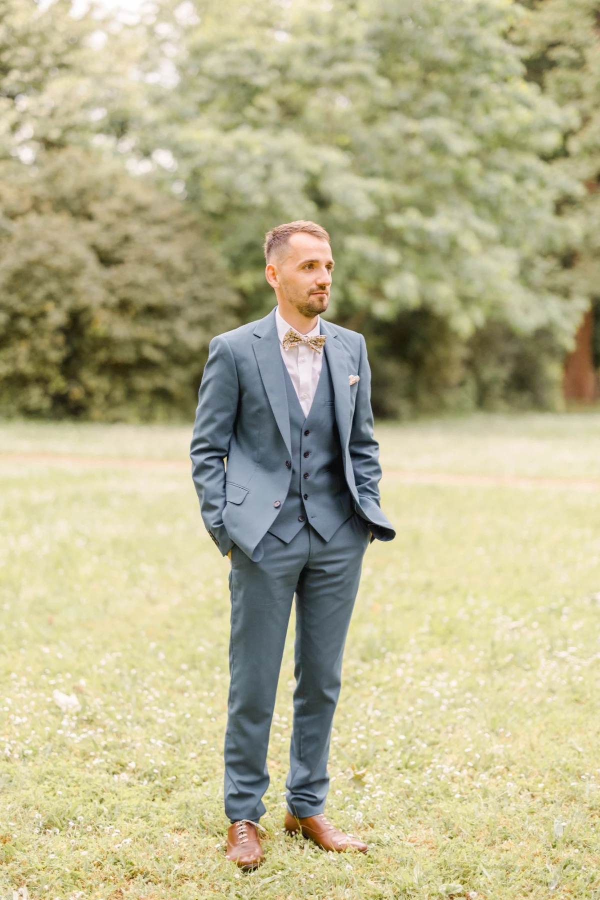 Marié en costume trois-pièces gris et nœud papillon se tenant dans un jardin verdoyant lors d'un mariage