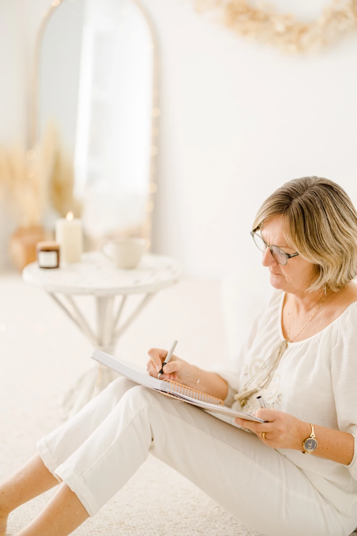 Femme blonde en tenue beige écrivant dans un carnet, assise près d'un miroir dans un intérieur lumineux