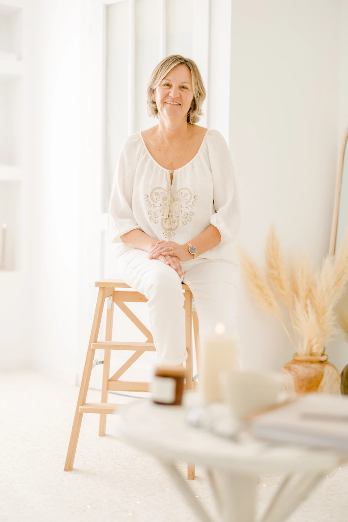 Portrait professionnel d'une femme souriante en tenue blanche brodée, assise sur un tabouret en bois