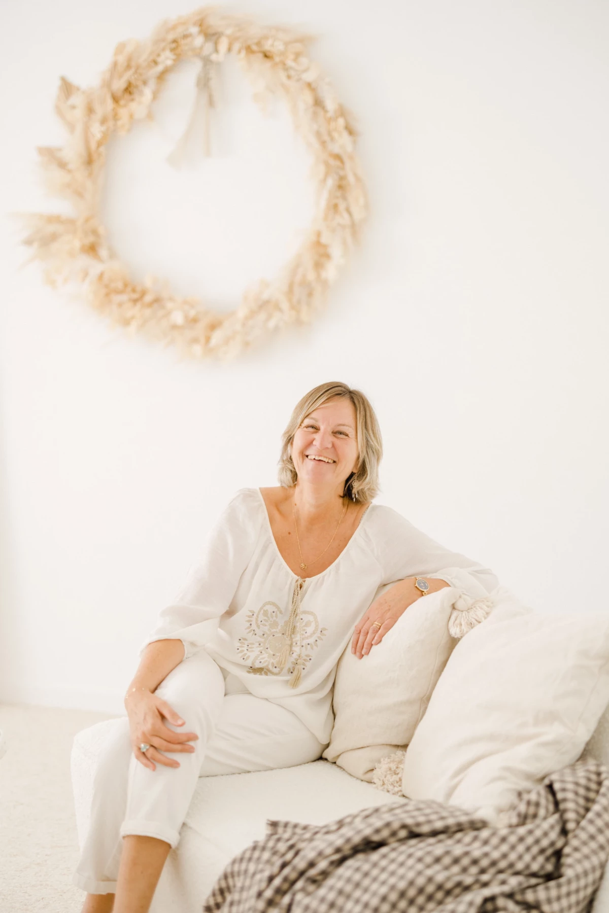 Portrait professionnel d'une femme souriante vêtue de blanc, assise sur un canapé beige dans un studio lumineux